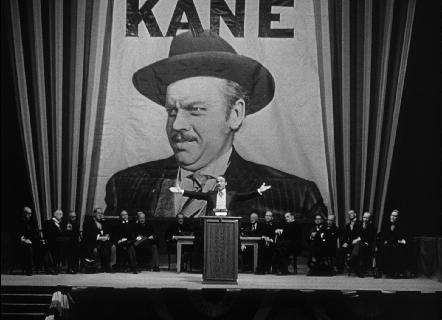Visuel Citizen Kane : un homme prononce un discours devant un immense portrait de lui-même, sur une estrade décorée de drapeaux.