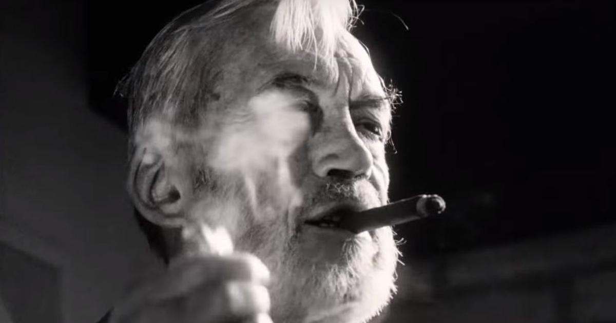 Plan en noir et blanc d’un homme âgé fumant un cigare, filmé en contre-plongée, avec la fumée flottant devant son visage.